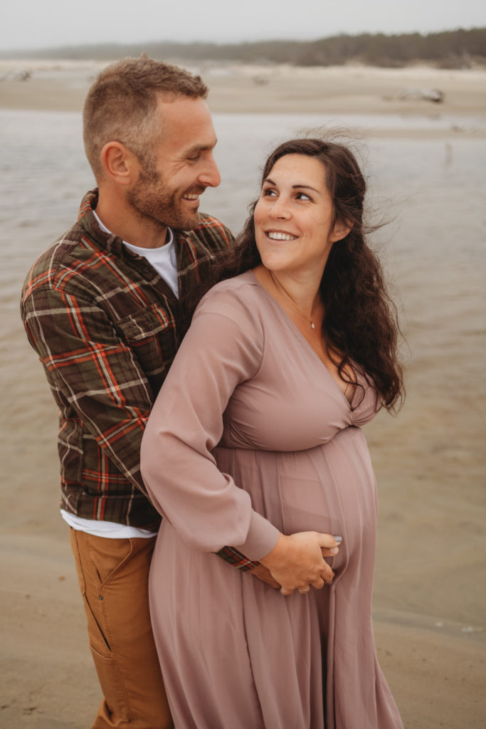 Wife smiling as she looks back at her husband on the Oregon Coast during a playful maternity session