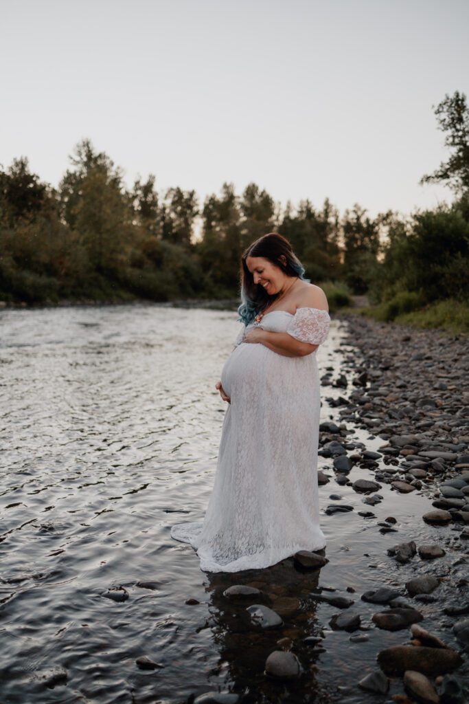 Pregnant woman in white lace dress looking down at baby bump during personalized family maternity session in Eugene Oregon