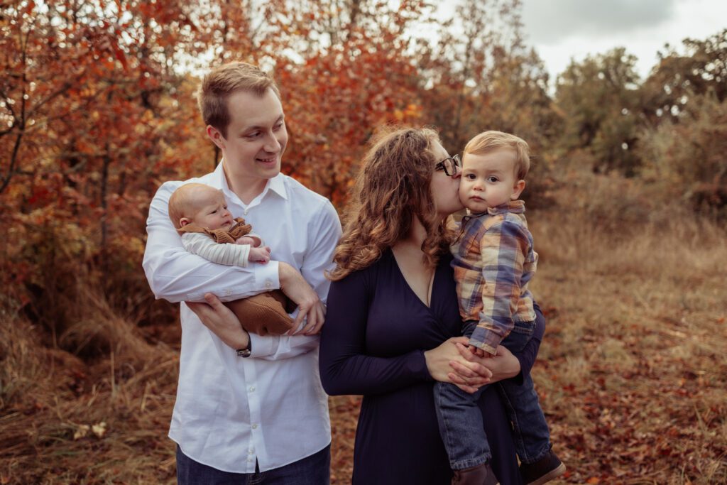 Fall family photos with family of 4, parenting holding newborn baby and kissing young child on cheek, at Dorris Ranch in Springfield Oregon