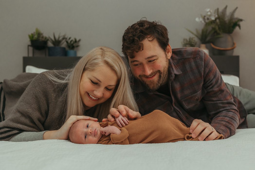 New parents smiling and laying on their bed admiring their newborn baby at 2 weeks old