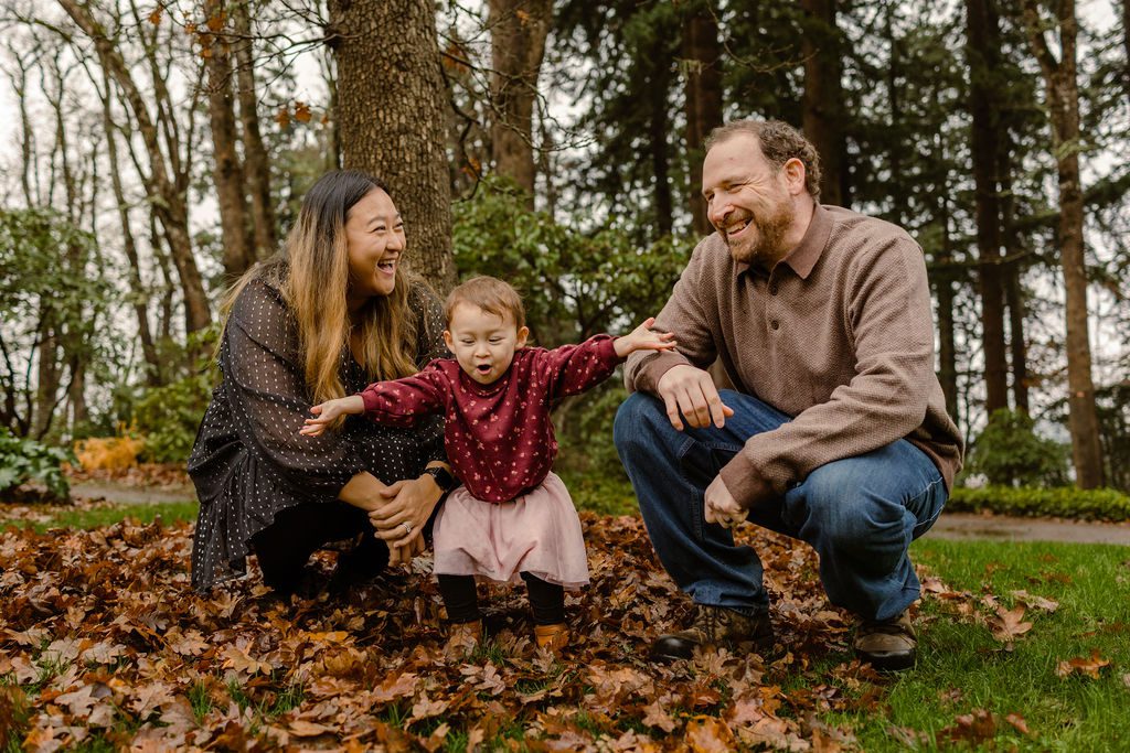 Young family laughing as two year old daughter throws leaves in the air during fall family photos at Hendricks Park in Eugene, Oregon 