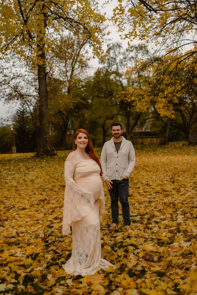 Vibrant fall maternity session at Alton Baker Park in Eugene, Oregon with husband standing slighting behind, admiring pregnant wife, and expectant mom holding belly smiling at the camera 