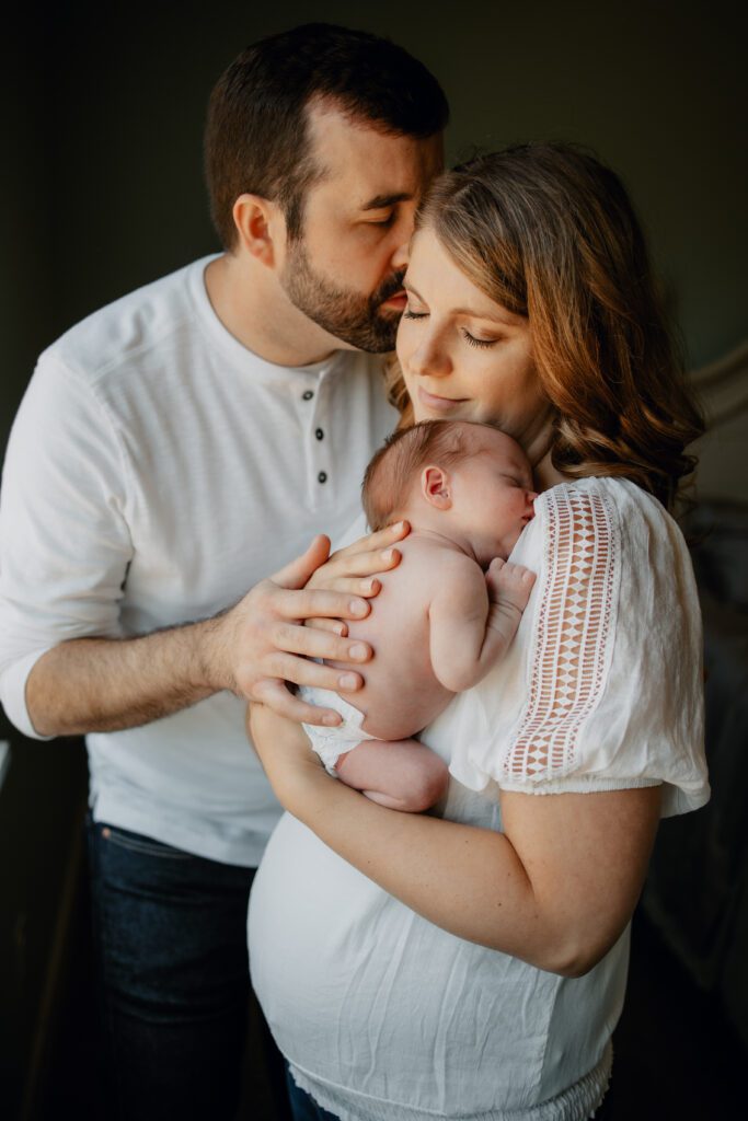 Dad in white Henley and jeans kissing mom in white lace top and jeans holding sleeping newborn baby in diaper during newborn photo session with Eugene Oregon newborn photographer Lux Marina Photography