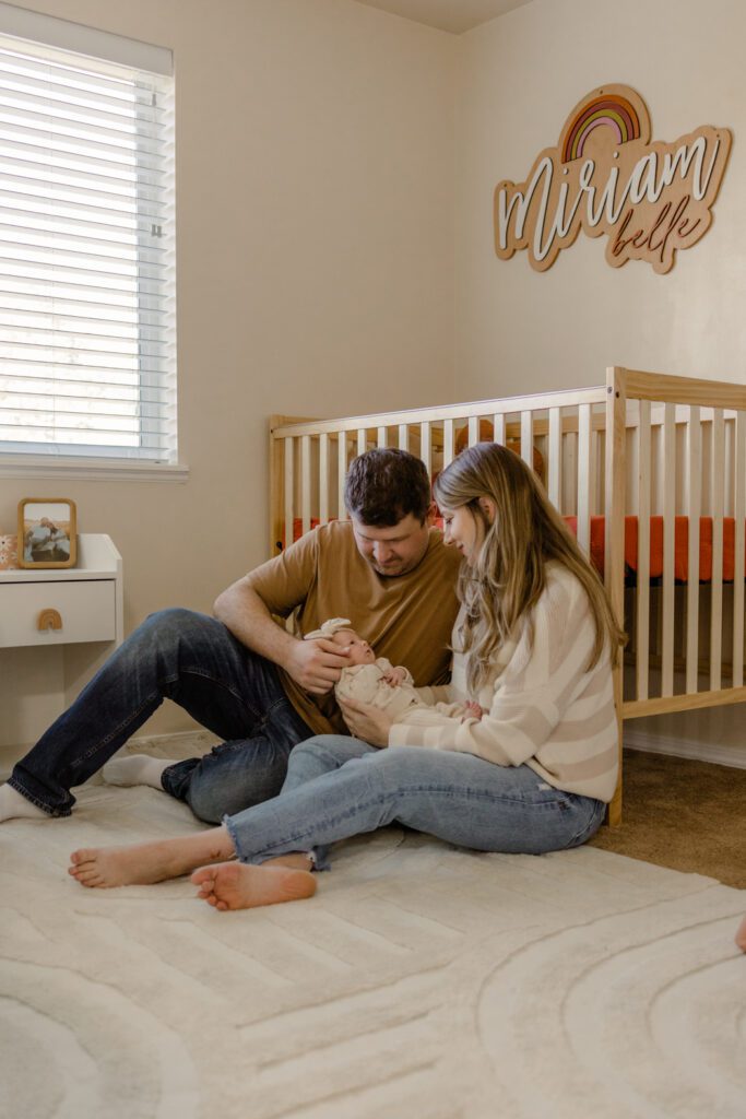 Father in jeans and brown shirt and mother in jeans with white and tan knit sweater lovingly looking at newborn baby wearing cream outfit during newborn photos