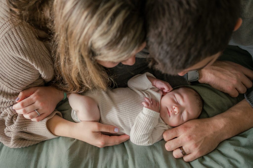 Mom wearing tan knit sweater and black leggings dad wearing gray sweater laying on bed with newborn baby wearing cream thermal onesie during loving newborn photography session