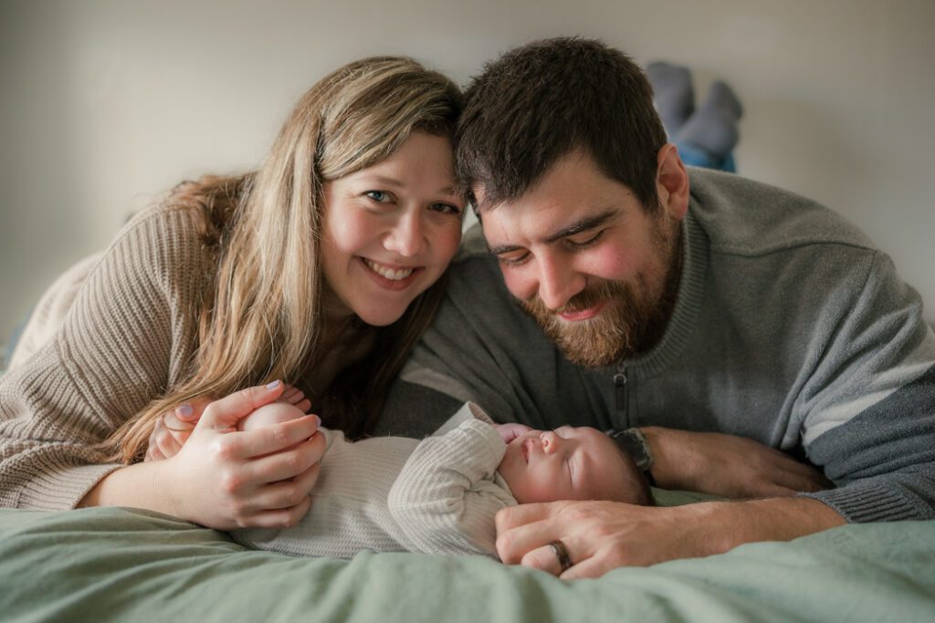 Parents and newborn baby laying on bed smile during in-home newborn session