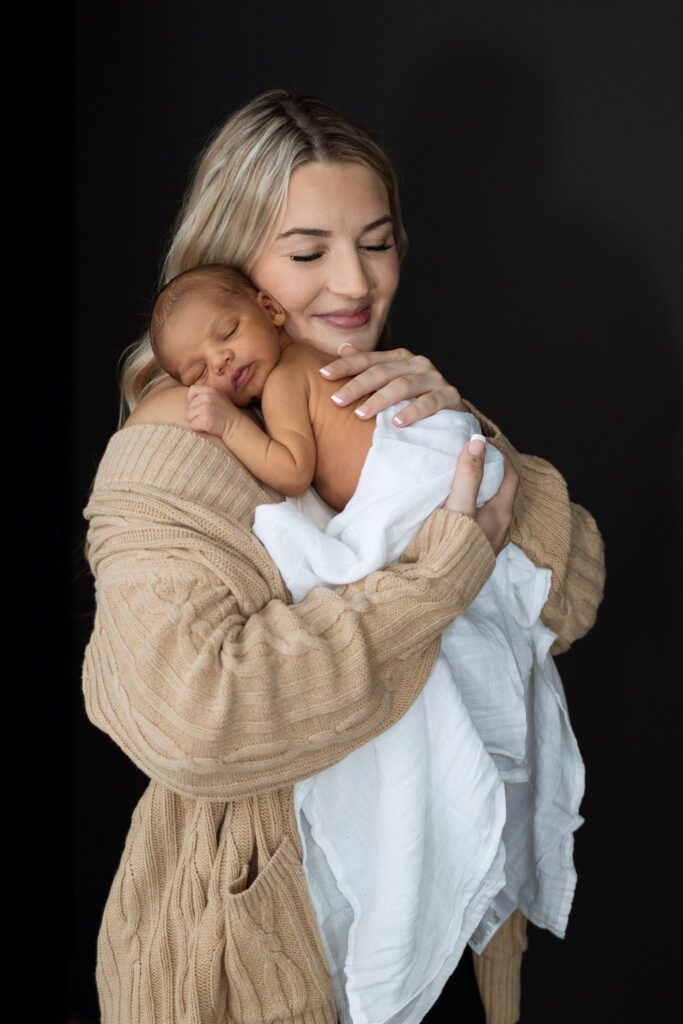 Mother in tan cardigan hugging sleeping newborn baby wearing white muslin blanket offering classic and neutral outfit ideas for newborn photo session