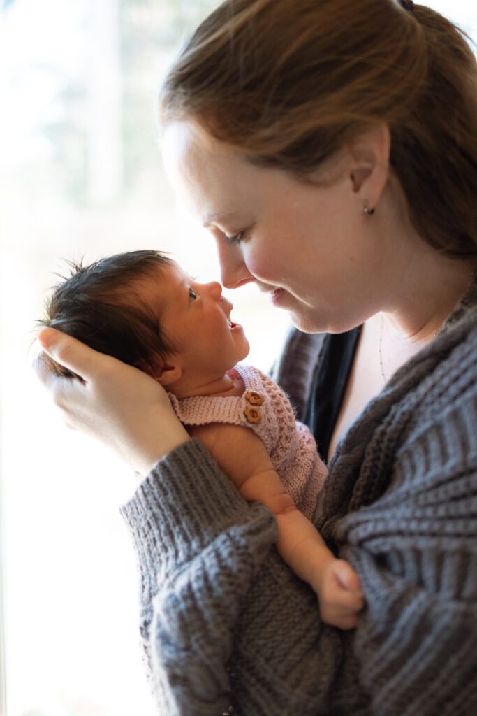 Mom wearing gray cardigan holding newborn baby wearing pink knit romper during in home newborn session with Eugene Oregon based newborn photographer Lux Marina Photography