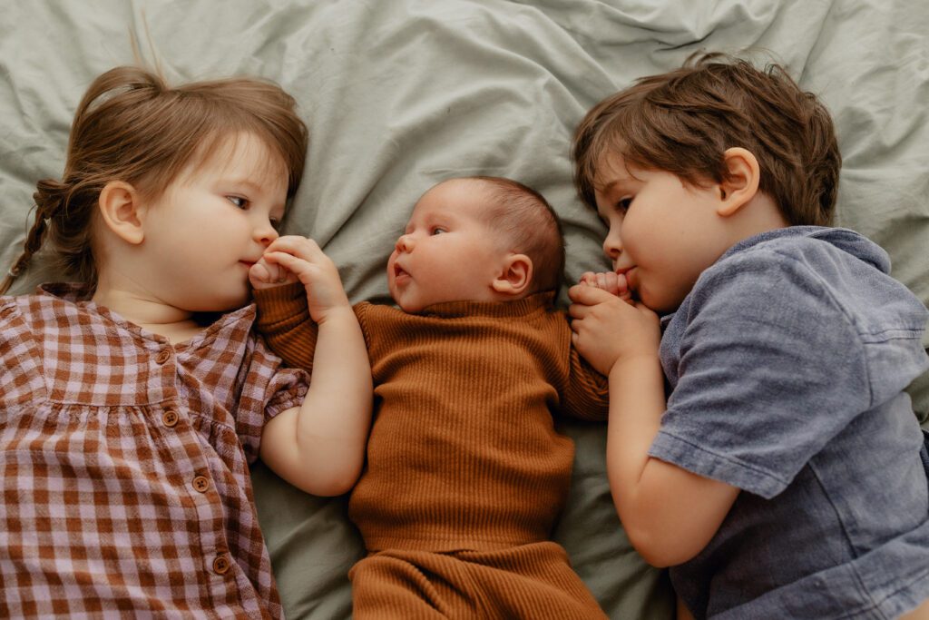Sister wearing brown and purple gingham dress brother wearing blue button down laying next to newborn sister wearing rust two piece outfit showing an example of siblings outfits of what to wear for newborn photo session