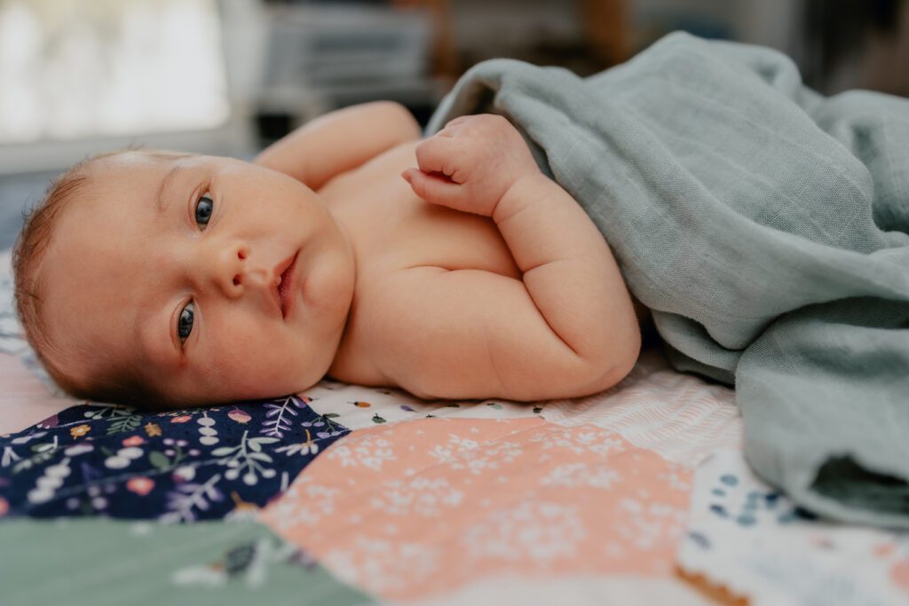 Newborn baby looking at camera laying on handmade floral quilt wearing blue muslin blanket showing personalized touches of what to wear for newborn photo session